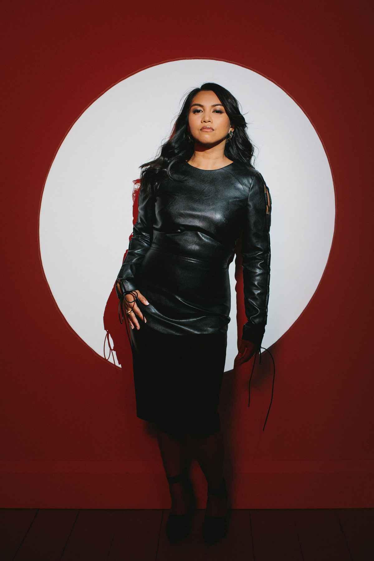 A creative portrait of a women in a black leather dress. She is standing in a white spotlight with red light shadows around her.