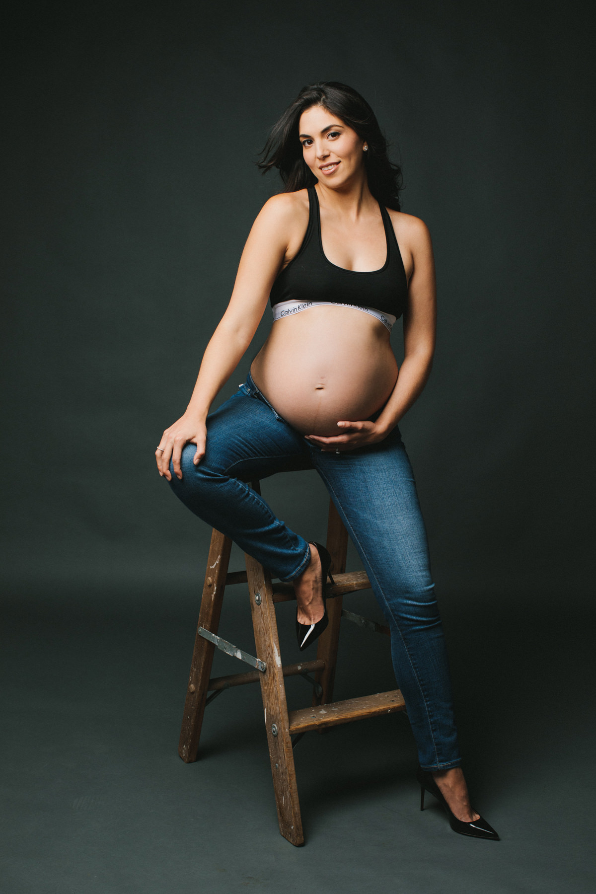 A modern maternity portrait of a mother to be. She is wearing black heals, denim and a black sports bra.