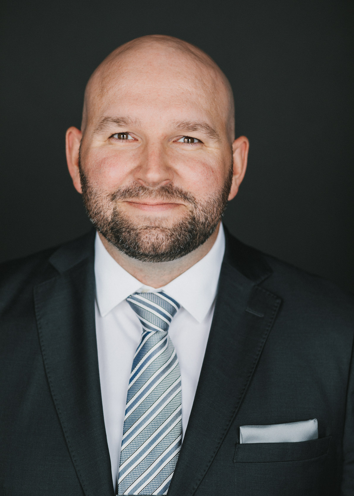 A studio headshot portrait of a male Lawyer in a black business suit with a white shirt and blue striped tie.