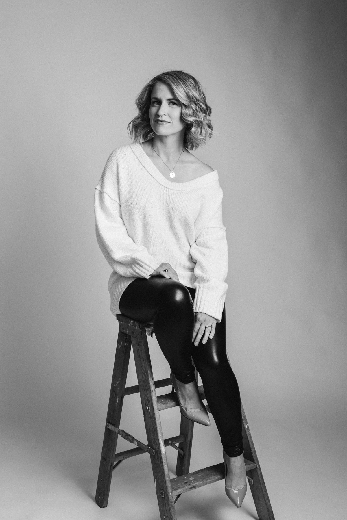 A black and White portrait of a women sitting on a wooden stepladder. This was photographed in a studio. The women is wearing a long white sweater and black leggings with high heel shoes.