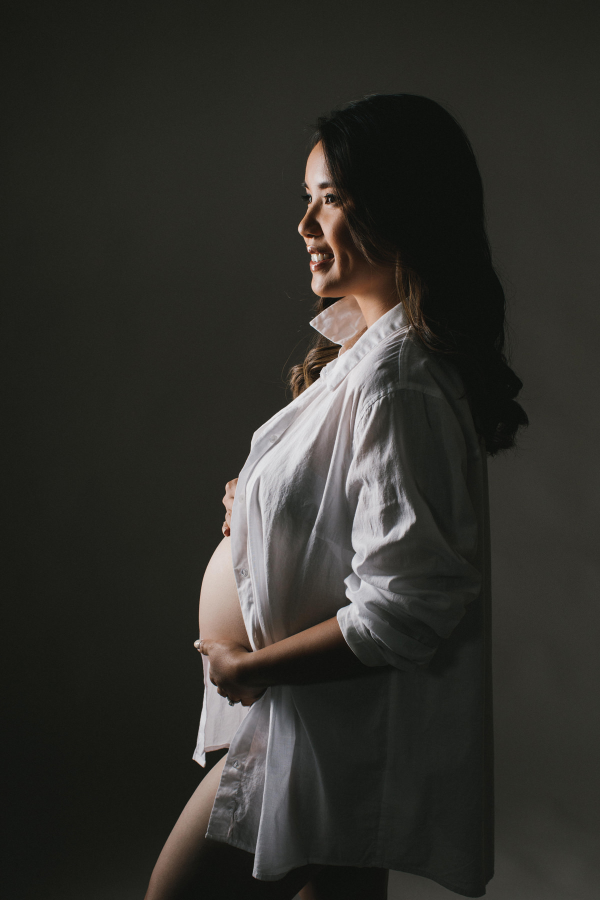 A maternity portrait created in studio of a woman in an oversized white dress shirt.