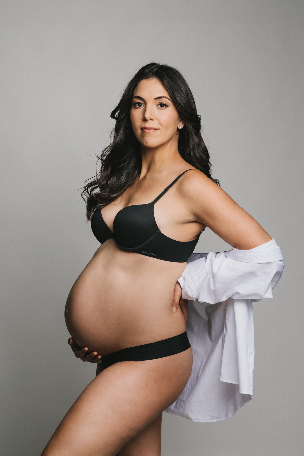 A maternity portrait of a beautiful expecting women. She is wearing a black underwear set and an oversized white dress shirt.