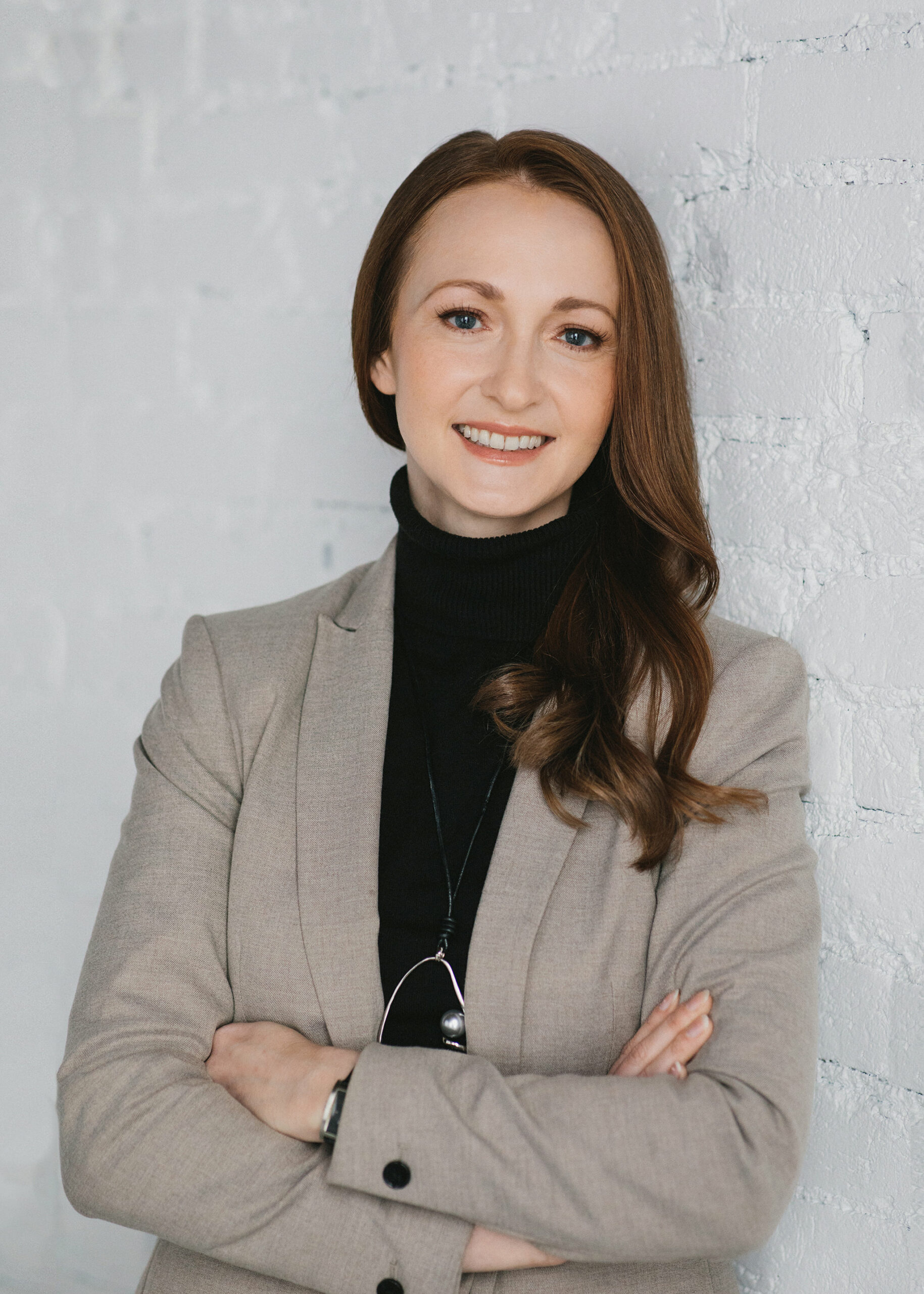 A business portrait of a women in a beige blazer and black turtleneck sweater. She is leaning again a white brick wall.